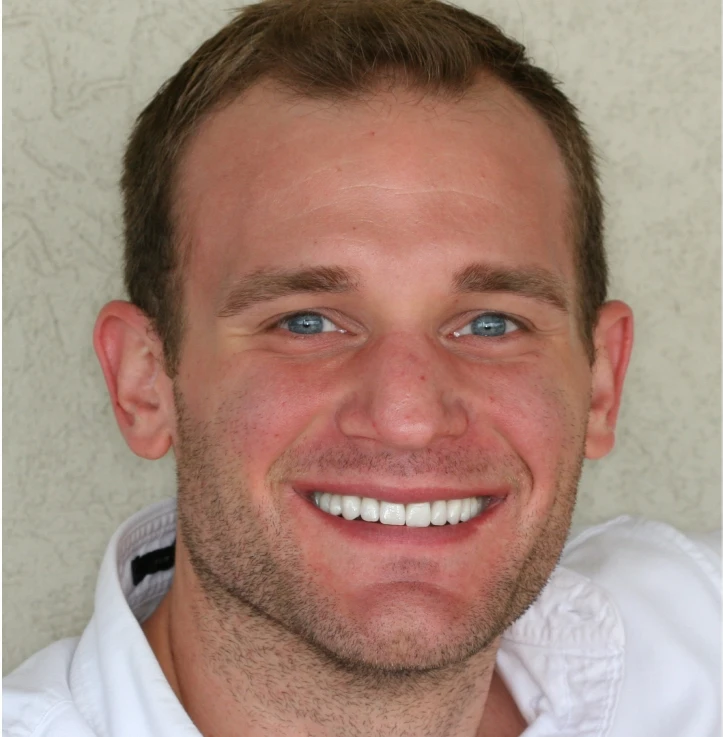 Close-up of a smiling man with short brown hair, blue eyes, light stubble, and wearing a white shirt.