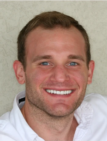 Smiling man with blue eyes, short brown hair, light stubble, wearing a white collared shirt.