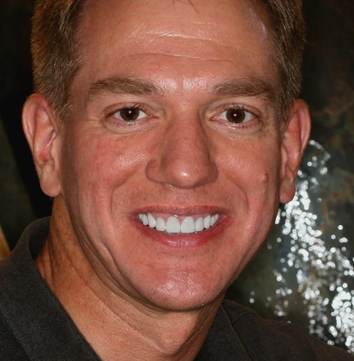 Close-up of a smiling man with short brown hair and brown eyes wearing a dark shirt.