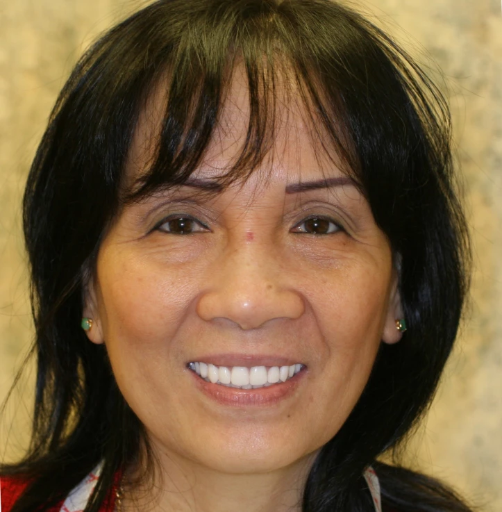 Smiling middle-aged woman with dark hair, wearing small green earrings, in front of a light brown patterned background.