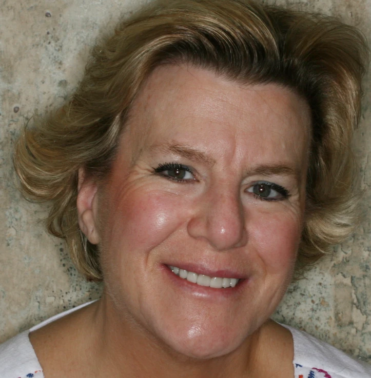 Smiling middle-aged woman with short blonde hair and light makeup against a textured wall background.