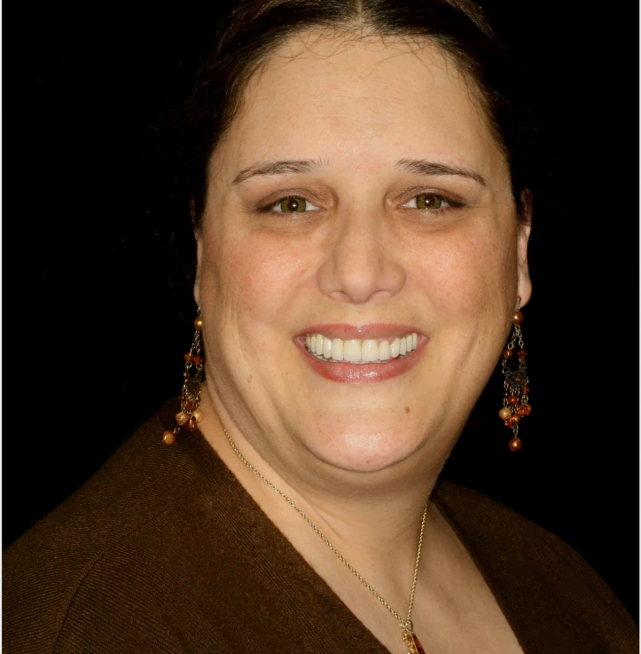 Smiling woman with brown eyes wearing dangling orange and gold earrings and a brown top against a black background.