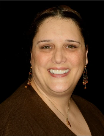 Smiling woman with brown hair wearing dangling earrings and a necklace against a black background.