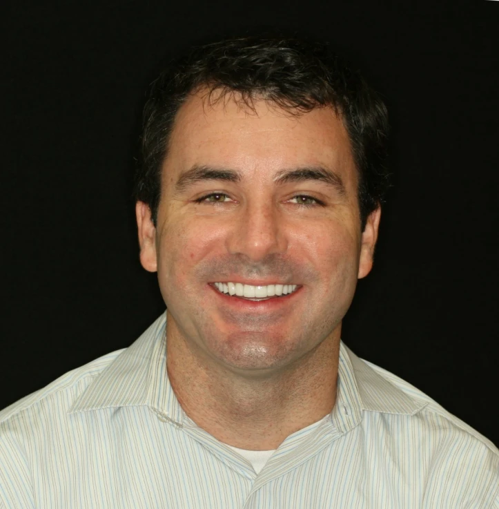 Smiling man with dark hair wearing a light striped button-up shirt against a black background.