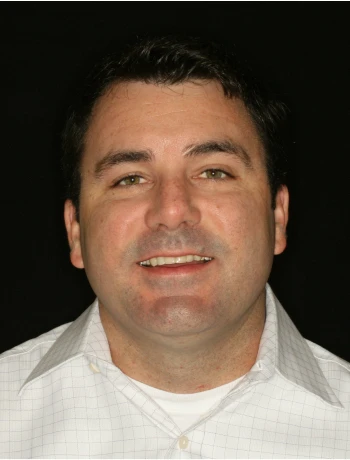 Smiling man with short dark hair wearing a white collared shirt against a black background.