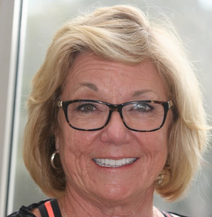 Smiling older woman with short blonde hair, wearing black-rimmed glasses and silver hoop earrings.