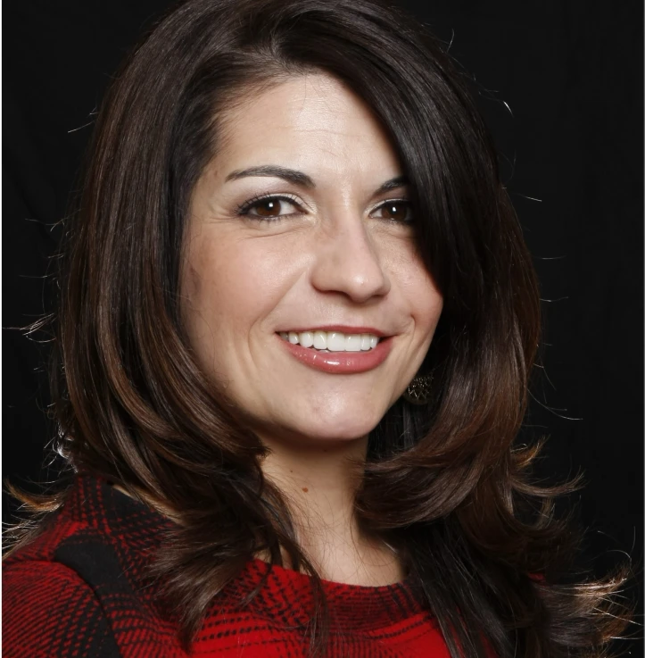 Smiling woman with dark brown hair wearing a red and black plaid top against a black background.