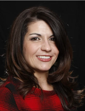 Smiling woman with dark brown hair wearing a red and black plaid top against a black background.