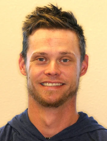 Smiling man with short, messy dark hair and light facial hair wearing a navy blue hoodie against a beige background.