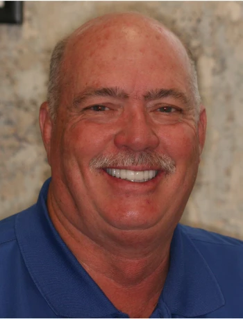 Smiling middle-aged man with a mustache wearing a blue collared shirt against a neutral background.