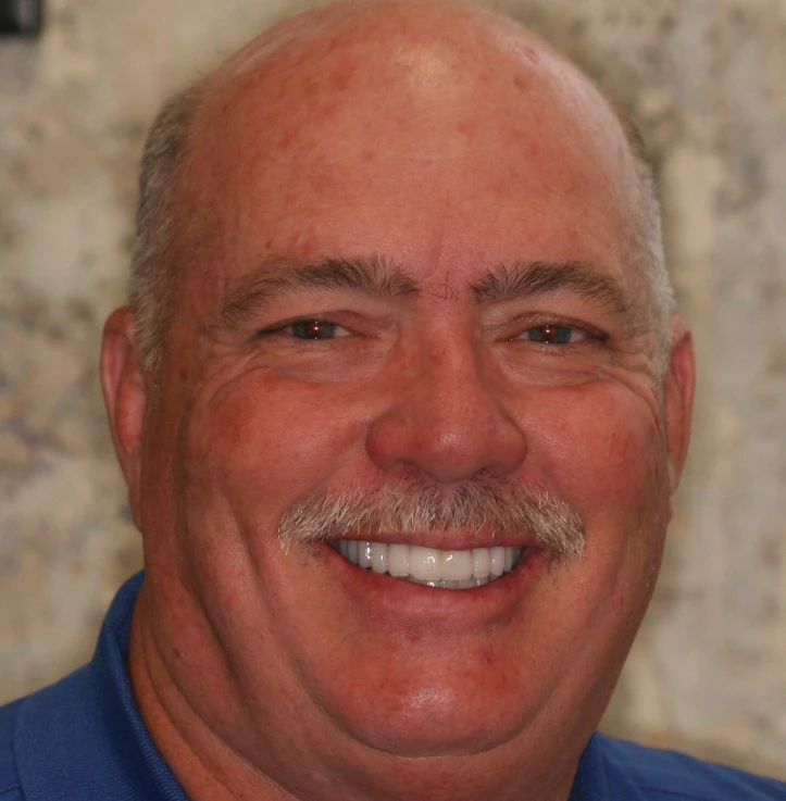 Smiling bald man with a gray mustache wearing a blue shirt.