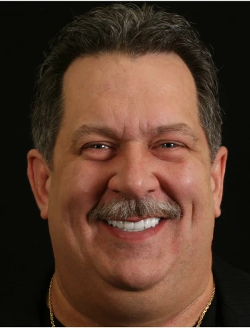 Smiling middle-aged man with a mustache and short dark hair against a black background.
