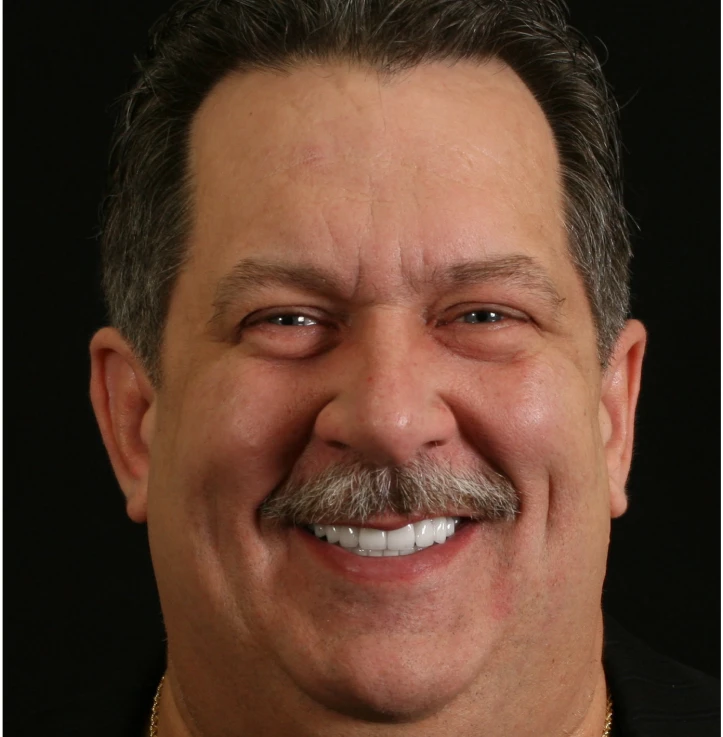 Middle-aged man with dark hair and mustache smiling against a black background.