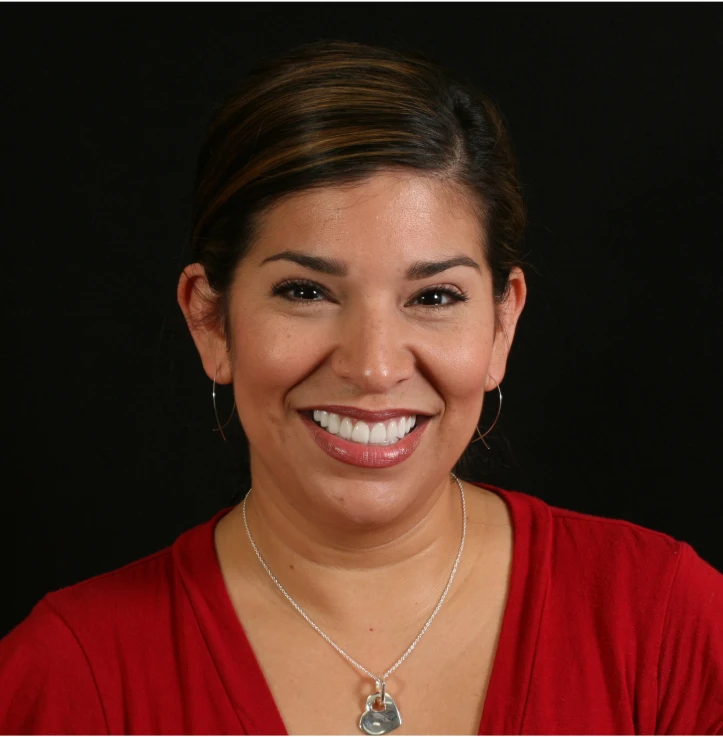 Smiling woman with dark hair, wearing a red top, hoop earrings, and a silver necklace with a heart pendant against a black background.