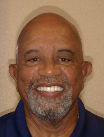 Smiling bald man with a gray beard wearing a navy blue shirt against a beige background.