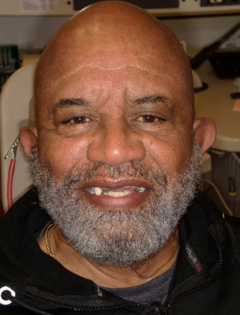 Smiling bald man with a graying beard wearing a black jacket indoors.