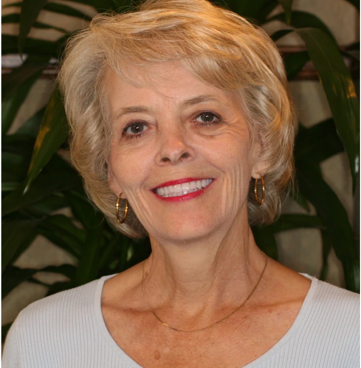Smiling older woman with short blonde hair, wearing gold hoop earrings and a light blue top.