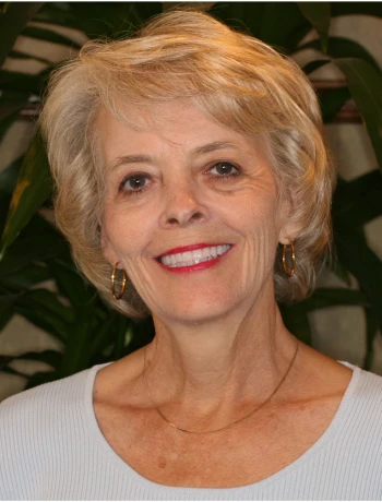 Smiling elderly woman with short blonde hair, wearing gold hoop earrings and a light blue top, in front of leafy green plants.