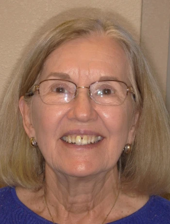 Smiling older woman with shoulder-length light blonde hair, glasses, and wearing a blue top.