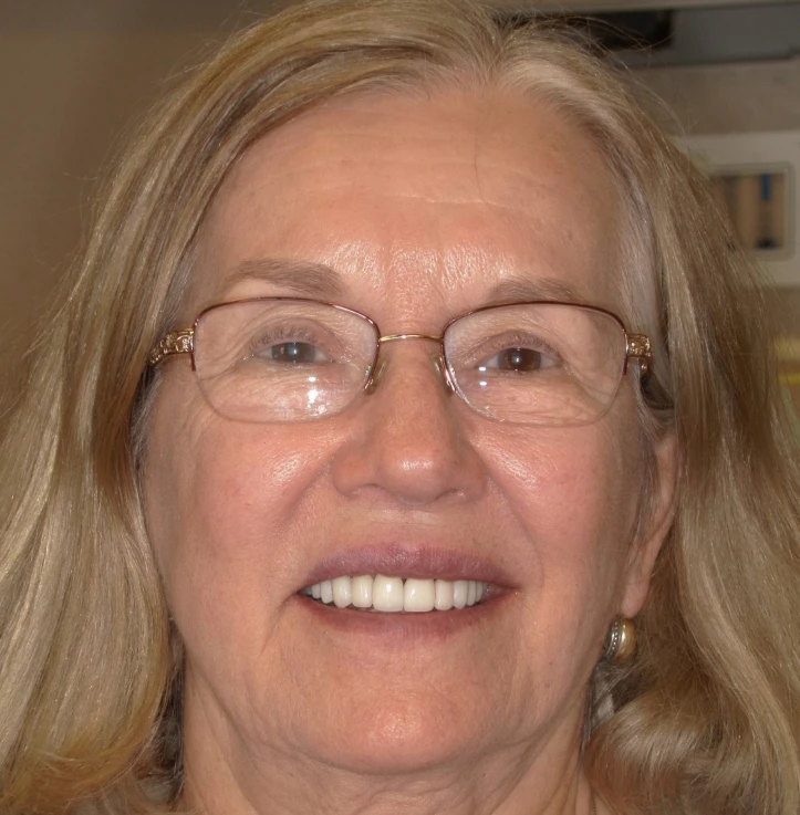 Smiling older woman with long blonde hair, glasses, and gold earrings.