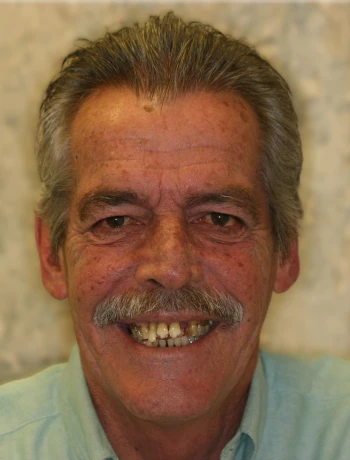 Smiling older man with gray hair, mustache, and missing front teeth, wearing a light blue shirt.
