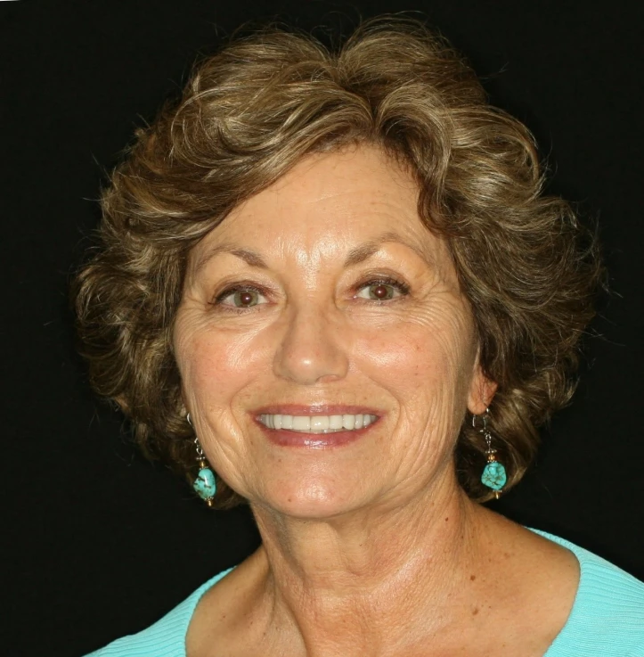 Smiling older woman with short curly hair wearing turquoise earrings and a light blue top against a black background.