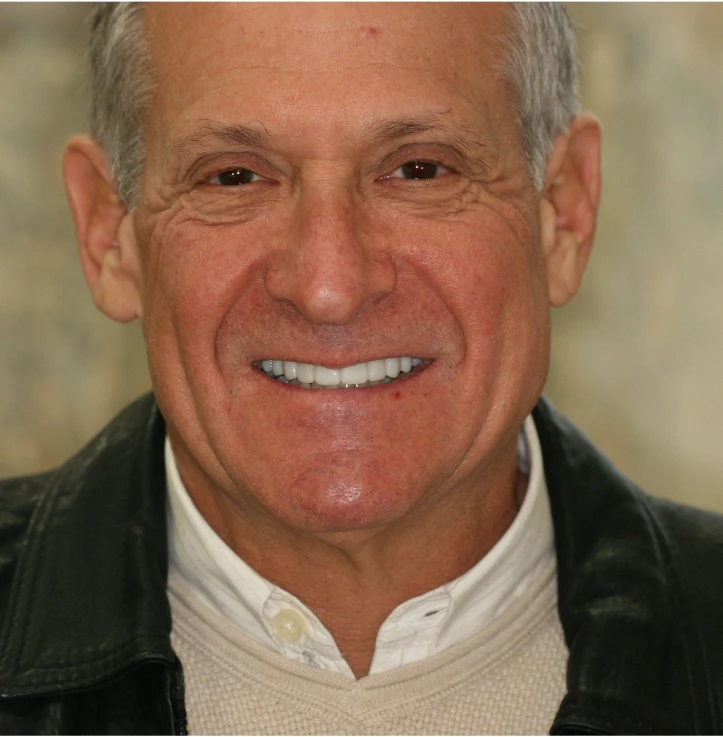 Smiling older man with gray hair wearing a white shirt, beige sweater, and black jacket.