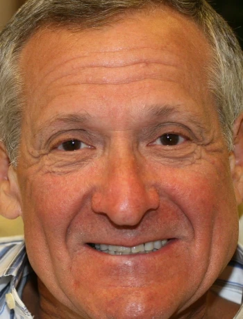 Close-up of an older man with gray hair smiling and wearing a checkered shirt.