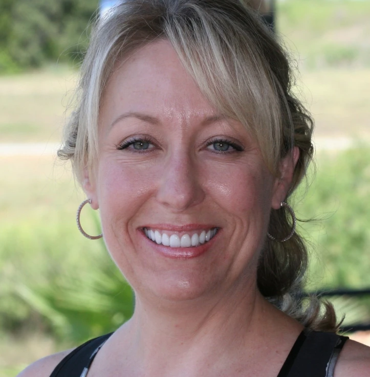 Smiling woman with blonde hair wearing hoop earrings and a sleeveless top, outdoors with blurred greenery in the background.