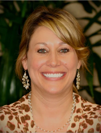 Smiling woman with blonde hair wearing pearl necklace, dangling earrings, and a leopard print top against a dark green background.