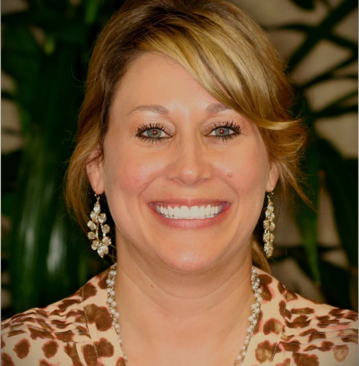 Smiling woman with blonde hair wearing dangling earrings, a pearl necklace, and a leopard print top, with blurred green plants in the background.