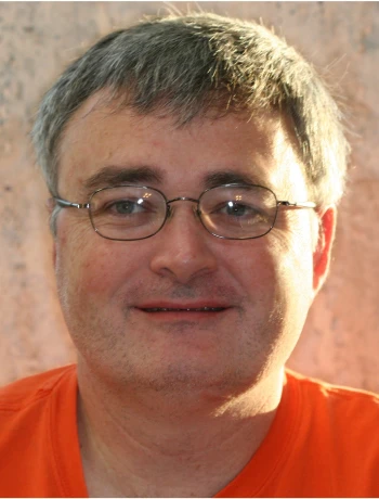 Middle-aged man with short gray hair and glasses smiling, wearing an orange shirt.