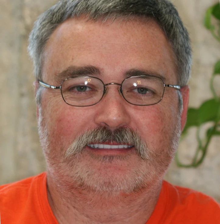 Close-up of a middle-aged man with gray hair, glasses, and a mustache, wearing an orange shirt.