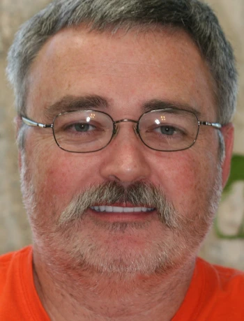 Close-up of a man with gray hair, beard, glasses, and a slight smile wearing an orange shirt.