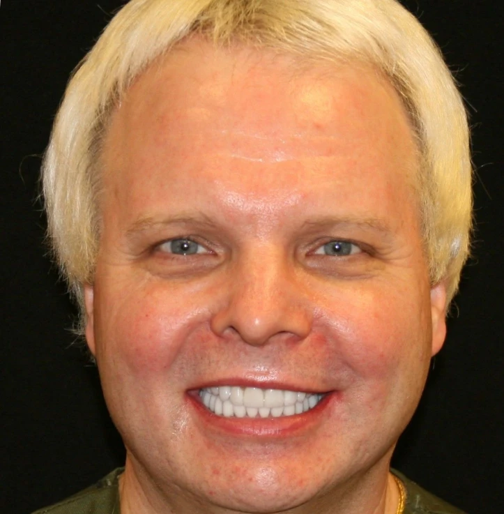 Smiling man with short blond hair and blue eyes against a black background.