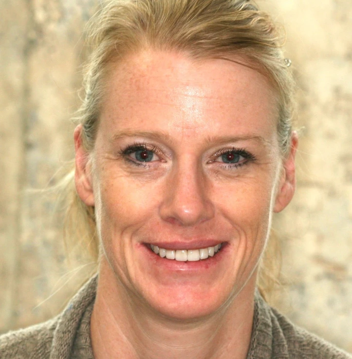 Close-up of a woman with light blonde hair pulled back, smiling at the camera against a blurred stone background.