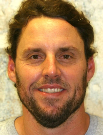 Smiling man with short brown hair, a beard, and a light gray shirt against a blurred background.