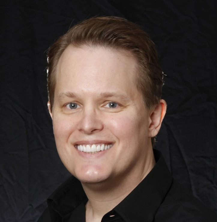 Smiling man with light skin and short light brown hair wearing a black shirt against a dark background.