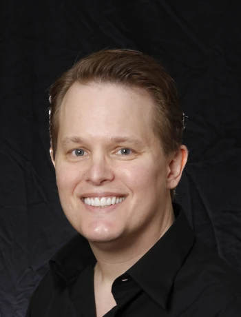 Smiling man with short light brown hair wearing a black shirt against a black background.
