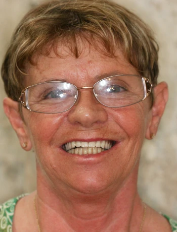 Smiling elderly woman with short hair, glasses, and small earrings against a blurred background.