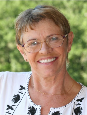 Smiling middle-aged woman with short brown hair, glasses, and a white blouse with black embroidery, standing outdoors with a green blurred background.