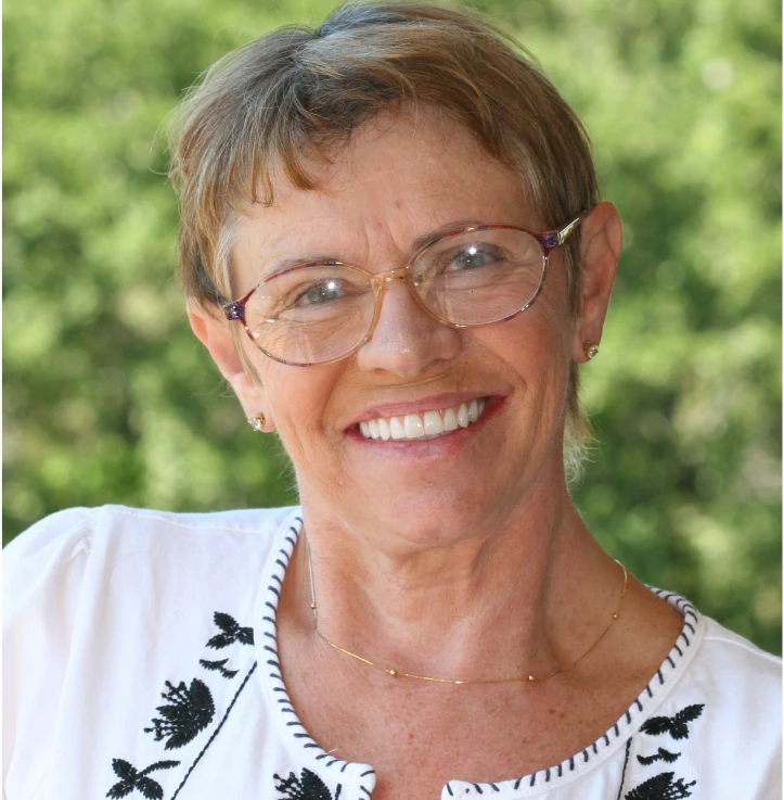 Smiling older woman with short light brown hair wearing glasses and a white top with black floral embroidery.