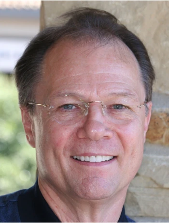 Smiling middle-aged man with glasses and short dark hair standing outdoors near a stone wall.