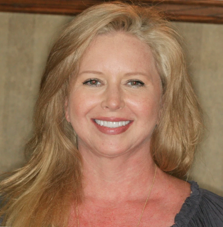 Smiling blonde woman with long hair wearing a dark top and gold necklace in front of a beige background.