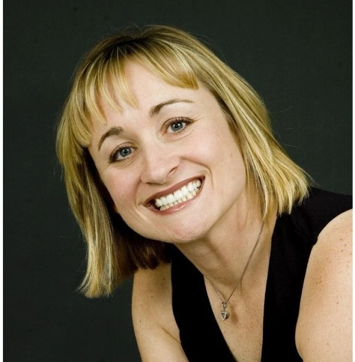Smiling woman with short blonde hair wearing a black sleeveless top and a pendant necklace against a dark background.