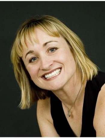 Smiling woman with short blonde hair wearing a black sleeveless top and a silver necklace against a dark background.