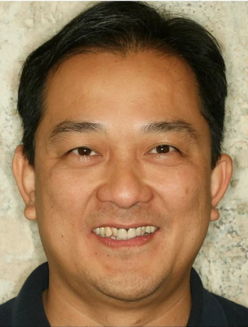 Close-up of a smiling middle-aged Asian man with short black hair against a light stone background.