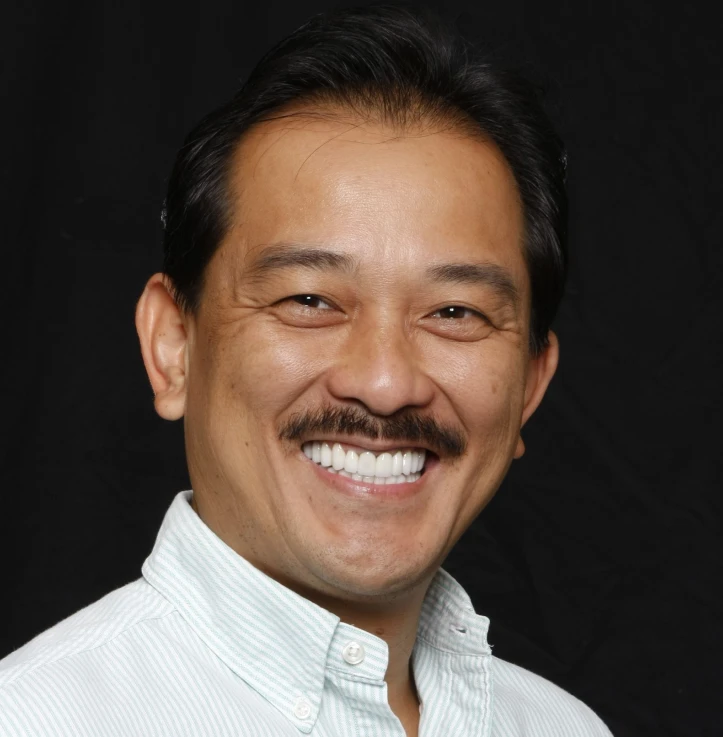 Smiling man with dark hair and a mustache wearing a light-colored button-up shirt against a black background.