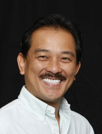 Smiling man with a mustache wearing a light striped button-up shirt against a black background.