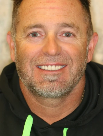 Close-up of a smiling middle-aged man with short hair, beard, and wearing a black hoodie with neon green drawstrings.